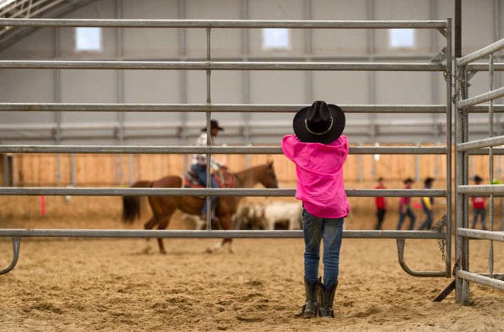 A young child wearing a cowboy hat stands outside a paddock watching a horseback rider.